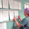 Person uprights documents in a softly curved aluminum document holder rail on a wall with multiple rows of papers, in a work environment resembling a hospital