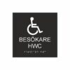 Panneau noir et blanc indiquant les toilettes pour visiteurs (HWC), avec un pictogramme représentant un fauteuil roulant ; le texte « Toilettes pour visiteurs (HWC) » figure sous le symbole et le braille en bas